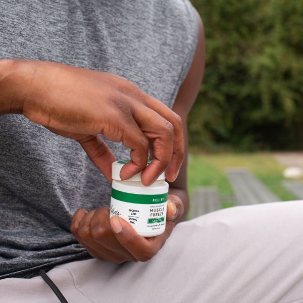 A person outdoors holding a jar of Mary's Relief Muscle Freeze, with their hand unscrewing the lid. The background shows a blurred green park setting.